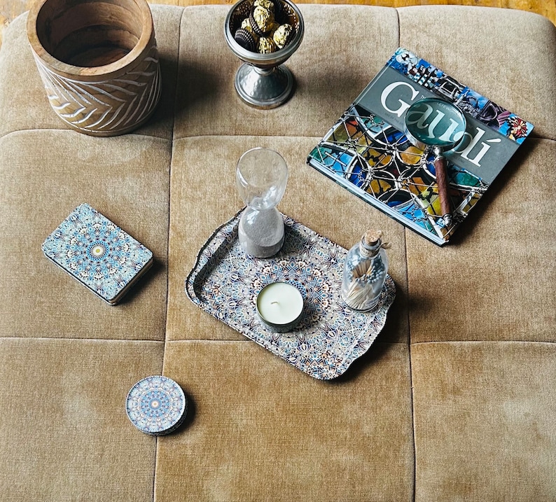 May include: A tray with a blue and white patterned design, featuring a small white candle in the center. The tray is on a beige surface with a wooden bowl, a glass hourglass, a book with the word "Gaudi" on the cover, and a magnifying glass.