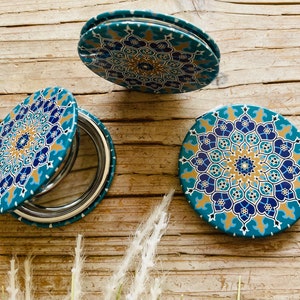 May include: Three round, compact mirrors with a blue, gold, and white geometric pattern. The mirrors are closed and feature a traditional design.