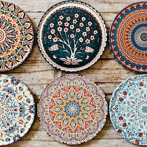 May include: Six decorative round plates with intricate, colourful patterns. The plates feature a variety of designs, including floral, geometric, and abstract motifs in shades of red, blue, orange, and green. The plates are arranged on a wooden surface.