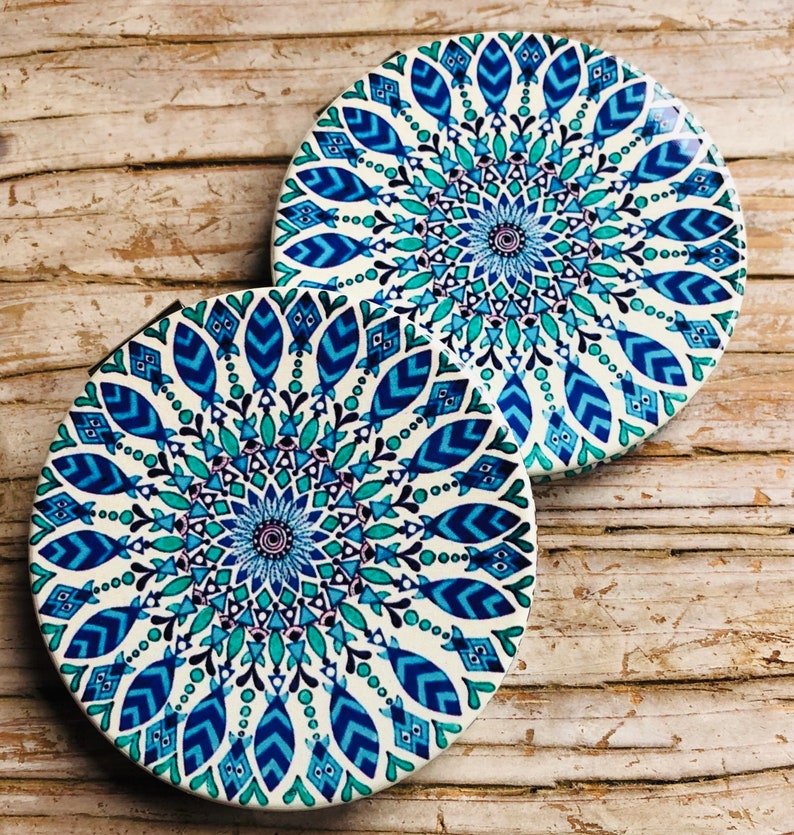 May include: Two round, white coasters with a blue and green mandala design. The coasters feature a circular pattern with intricate details.
