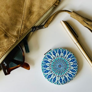 May include: A brown suede zippered pouch with a tassel, a pair of sunglasses, a black eyeliner, and a round compact mirror with a blue and white mandala design.