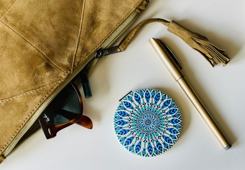 May include: A brown suede zippered pouch with a tassel, a pair of sunglasses, a black eyeliner, and a round compact mirror with a blue and white mandala design.