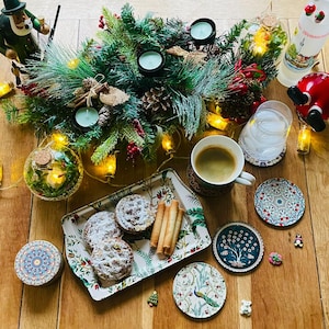 May include: A wooden table setting with a festive Christmas theme. A tray with three pastries, a glass of water, a cup of coffee, and a variety of coasters with intricate designs. The table is decorated with a green garland with lights, candles, and a small wooden figurine.