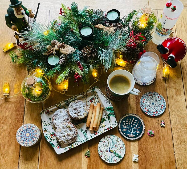 May include: A wooden table setting with a festive Christmas theme. A tray with three pastries, a glass of water, a cup of coffee, and a variety of coasters with intricate designs. The table is decorated with a green garland with lights, candles, and a small wooden figurine.