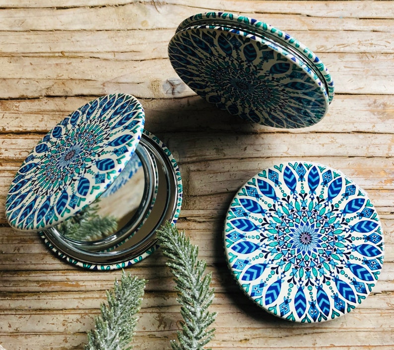 May include: Three round compact mirrors with a blue and white mandala design. The mirrors are closed and feature a silver reflective surface.