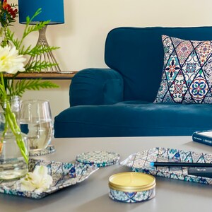 May include: A teal blue armchair with a colorful patterned pillow. A glass of water, a vase of white flowers, and a candle are on a table in front of the chair. There are also several decorative trays with a similar pattern to the pillow.