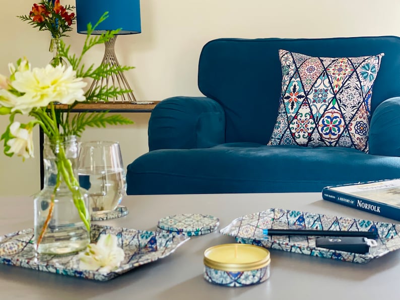 May include: A teal blue armchair with a colorful patterned pillow. A glass of water, a vase of white flowers, and a candle are on a table in front of the chair. There are also several decorative trays with a similar pattern to the pillow.