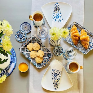 May include: A table setting with blue and white patterned plates, coasters, and napkins. There are also croissants, cookies, and a candle on the table.