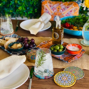 May include: A table setting with a variety of snacks and drinks, including olives, grapes, bread, and a glass of water. The table is decorated with colorful coasters and a patterned tablecloth.