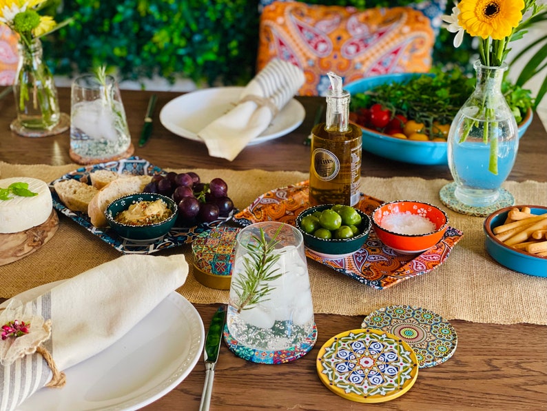 May include: A table setting with a variety of snacks and drinks, including olives, grapes, bread, and a glass of water. The table is decorated with colorful coasters and a patterned tablecloth.