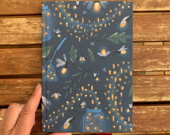 May include: A dark blue notebook with a pattern of fireflies and jars of fireflies. The fireflies are glowing yellow and the jars are clear glass.