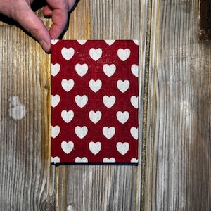 May include: A small, rectangular fabric item featuring a red backdrop adorned with a pattern of white hearts. The item is held by a hand against a wooden background.