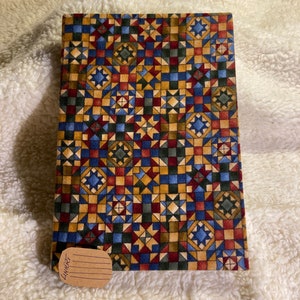 Könnte beinhalten: Ein brauner, blauer, roter und goldener Stoff-Notizbuchumschlag mit geometrischem Muster. Der Umschlag hat ein kleines braunes Papieretikett mit dem Wort "LINERT" darauf.