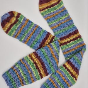 May include: Hand-knitted leg warmers in a striped pattern. The warmers feature bands of blue, green, purple, yellow, and maroon. The leg warmers are made of a soft, textured yarn and are designed to keep legs warm.