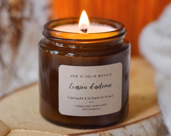 May include: A lit brown glass jar candle with a white label. The label reads "UNE SI JOLIE BOUGIE Evasion d'automne Fabriquée à la main en Alsace CRÉATION PARFUMÉE ARTISANALE". The candle is on a wooden surface.