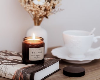 Puede incluir: Un tarro de vela de vidrio marrón con una etiqueta blanca que dice "Une So Jolie Journee" y "The Love of Coffee". La vela está encendida y la llama es visible. La vela está sentada sobre un libro con un fondo blanco.