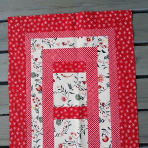 Könnte beinhalten: Ein roter und weißer Tischläufer mit einem Blumenmuster. Der Läufer hat einen roten Rand mit weißen Punkten.