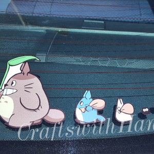May include: A set of five car decals featuring characters from the animated film My Neighbor Totoro. The decals include Totoro, a small Totoro, a catbus, and two soot sprites. The decals are black, brown, blue, and white. The decals are on a black car window. The text "Crafts with Hana" is visible in the bottom right corner of the image.