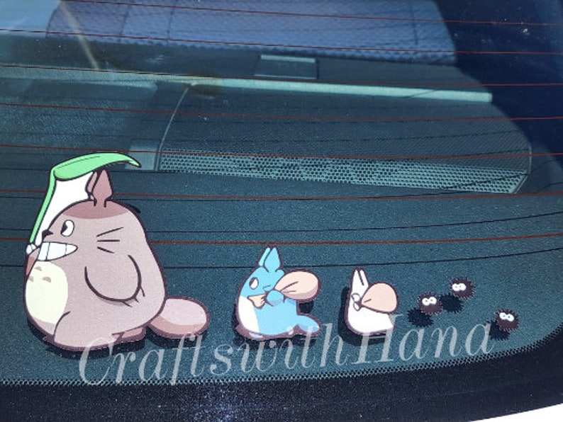 May include: A set of five car decals featuring characters from the animated film My Neighbor Totoro. The decals include Totoro, a small Totoro, a catbus, and two soot sprites. The decals are black, brown, blue, and white. The decals are on a black car window. The text "Crafts with Hana" is visible in the bottom right corner of the image.