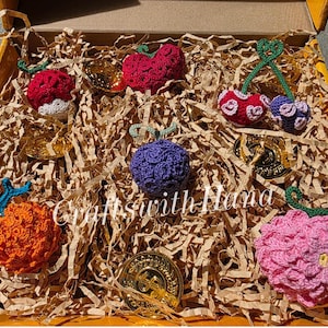 May include: A collection of colorful, crocheted fruit ornaments, including cherries, apples, and a purple berry, nestled in a box filled with shredded paper. The ornaments are in shades of red, pink, orange, and purple, with green stems.