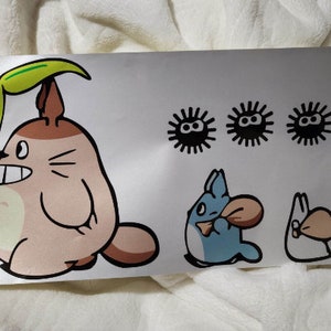 以下が含まれることがあります： となりのトトロのアニメ映画のキャラクターが描かれたステッカーシート。シートには、葉っぱを持ったトトロ、小さなトトロ、青い猫のような生き物、そして黒と白の煤けた精霊が3体含まれています。