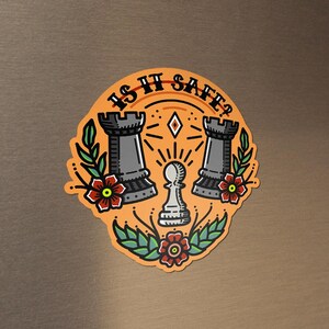 May include: A round, orange magnet with a chess-themed design. The magnet features two gray rook chess pieces, a white pawn, and the text "IS IT SAFE?" in a stylized font. Red flowers and green leaves surround the chess pieces.