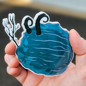 May include: A blue and white sticker with a pair of black horns and a white branch. The sticker is shaped like a rock.
