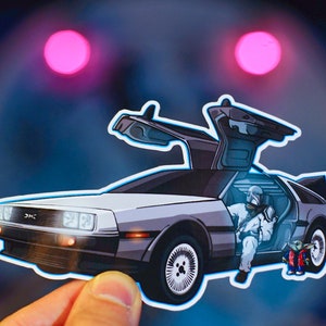 May include: A silver DeLorean time machine with the doors open. A figure in a white suit is sitting in the driver's seat. A small green figure is standing next to the car.