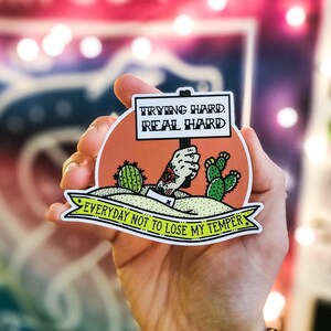 Op de afbeelding: Een sticker met een cartoonhand die een bord vasthoudt waarop staat "Trying hard, real hard. Everyday not to lose my temper." De hand draagt een armband en heeft een tatoeage. De sticker is op een perzikkleurige achtergrond met groene cactussen.
