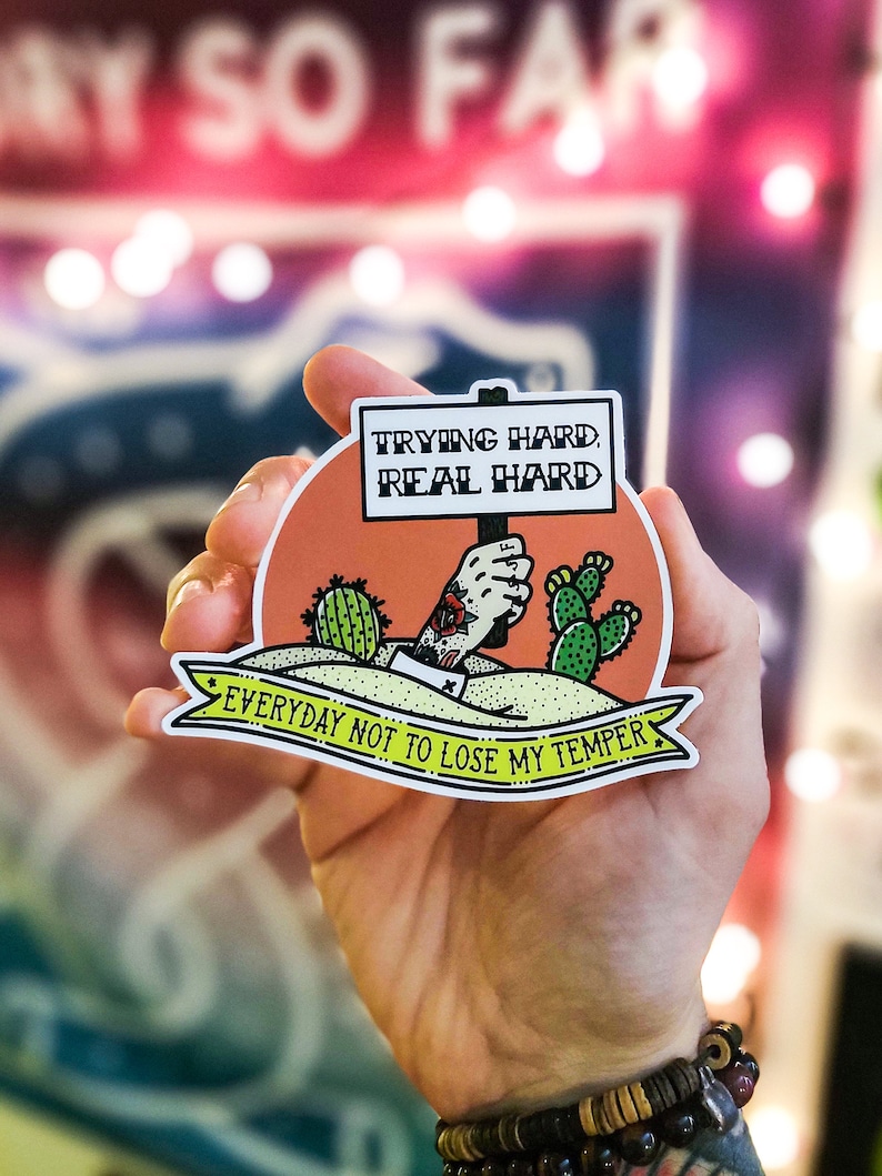 Op de afbeelding: Een sticker met een cartoonhand die een bord vasthoudt waarop staat "Trying hard, real hard. Everyday not to lose my temper." De hand draagt een armband en heeft een tatoeage. De sticker is op een perzikkleurige achtergrond met groene cactussen.