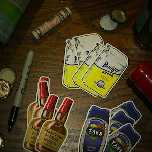 May include: Four vintage-style stickers on a wooden surface. The stickers feature various products, including a bottle of liquor, a bag of sugar, and a bottle of soda. The stickers are yellow, blue, and red.