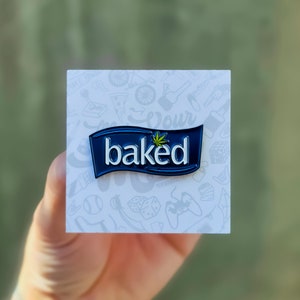 May include: A blue and white enamel pin with the word "baked" and a green marijuana leaf. The pin is on a white card with a variety of black and white illustrations.