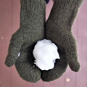 Puede incluir: Un par de guantes de lana verde oscuro sosteniendo una pequeña bola de nieve. Los guantes están hechos de una tela gruesa y texturizada.