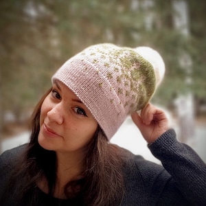 May include: A woman wearing a pink and green knitted hat with a geometric pattern. The hat is pulled down over her ears and she is looking at the camera.