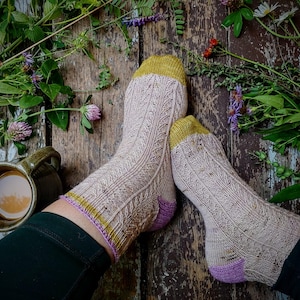May include: A pair of knitted socks with a beige background and a yellow and pink trim. The socks are on a wooden surface with green plants and a mug of coffee.
