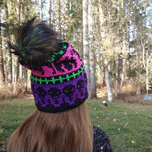 May include: A black knit hat with a pink and green patterned design featuring skulls and crossbones. The hat has a black faux fur pom pom on top.