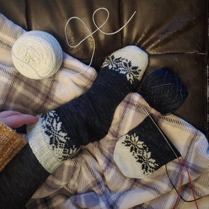 May include: A pair of blue and white knitted socks with a snowflake pattern. The socks are being knitted with white yarn and a black yarn ball.