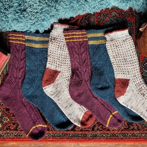 Könnte beinhalten: Fünf Paar bunte Stricksocken mit verschiedenen Mustern und Farben, darunter Burgunderrot, Teal und Weiß. Die Socken haben einen goldenen und gelben Streifen oben.