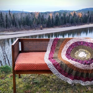 May include: A wooden bench with a red cushion and a woven backrest is draped with a circular, multi-colored knitted blanket. The blanket features concentric circles of brown, purple, and green. The scene is set outdoors with a river and trees in the background.