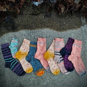 May include: A collection of nine colorful knitted socks in various patterns and colors, including pink, purple, blue, yellow, and white. The socks are laid out on a gray concrete surface.