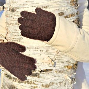 May include: Dark brown knitted gloves. The gloves are a solid color and appear to be made of a soft, warm material. The gloves are being held around a birch tree trunk. The person is wearing a cream-colored coat.