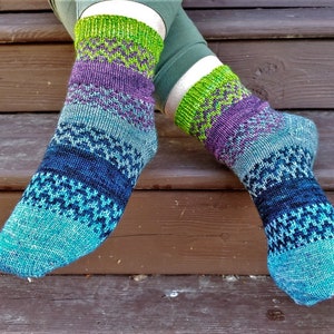 Puede incluir: Un par de calcetines de punto con un patrón geométrico. Los calcetines presentan bandas de color turquesa, azul, morado y verde. Se llevan en las piernas de una persona, con los pies apoyados en una superficie de madera.