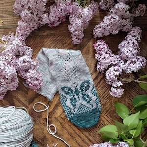 May include: A pair of knitted socks with a butterfly design in shades of teal and purple, surrounded by lilac blossoms.