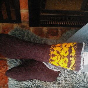 Könnte beinhalten: Ein Paar braune Socken mit gelben und orangenen Mustern. Die Socken werden an den Füßen einer Person getragen und liegen auf einer flauschigen grauen Oberfläche.