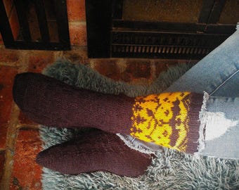 Peut inclure: Une paire de chaussettes marrons avec des motifs jaunes et orange. Les chaussettes sont portées aux pieds d'une personne et reposent sur une surface grise et duveteuse.