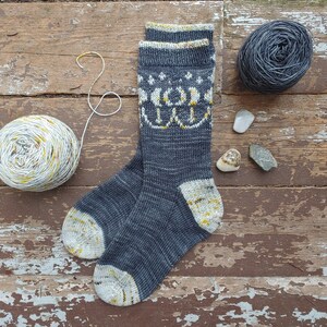 Puede incluir: Un par de calcetines de punto grises con un patrón geométrico blanco y amarillo. Los calcetines están sobre una superficie de madera.
