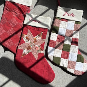 Op de afbeelding: Drie handgemaakte kerstsokken in rood, wit en groen. De namen "Mark", "Annie" en "Jude" zijn in het rood geborduurd. De sokken hebben gewatteerde en patchwork ontwerpen, met een sterpatroon op één.