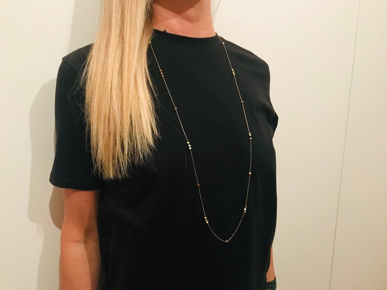 Op de afbeelding: Een lange gouden ketting met kleine, ronde, gouden kralen. De ketting wordt gedragen door een persoon met lang blond haar en een zwart T-shirt.