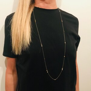 Op de afbeelding: Een lange, delicate gouden ketting met kleine, ronde, gouden kralen. De ketting wordt gedragen door een persoon met lang blond haar en een zwart T-shirt.
