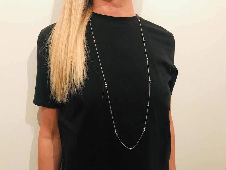 Op de afbeelding: Een lange, delicate gouden ketting met kleine, ronde, gouden kralen. De ketting wordt gedragen door een persoon met lang blond haar en een zwart T-shirt.